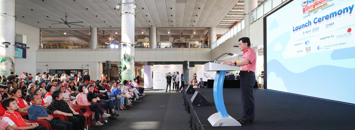 Man at podium speaks to audience. Screen reads "Launch Ceremony" and lists Singapore partners.